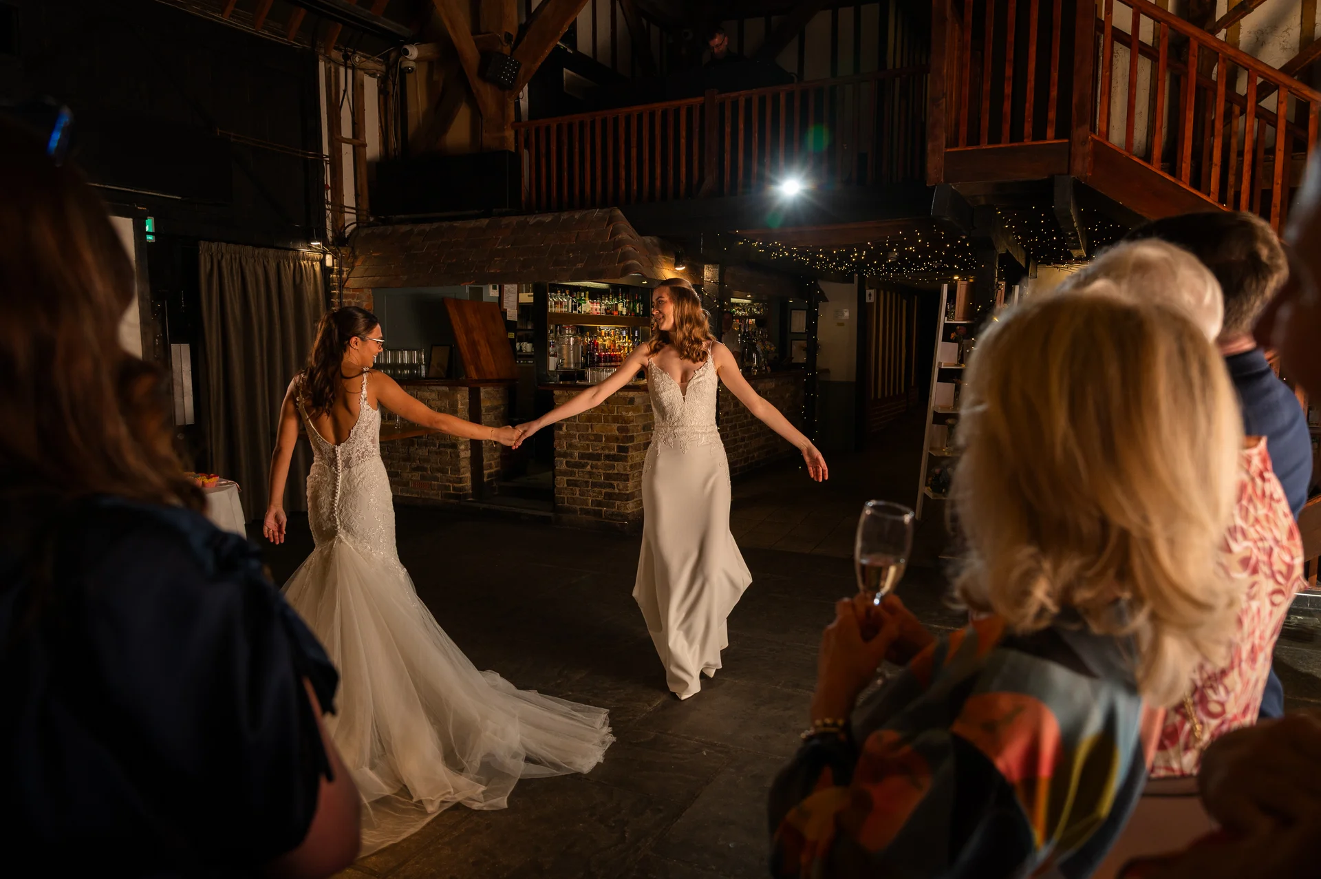 Two brides share a joyful first dance together holding hands inside a rustic barn venue illuminated by string lights while guests watch on.