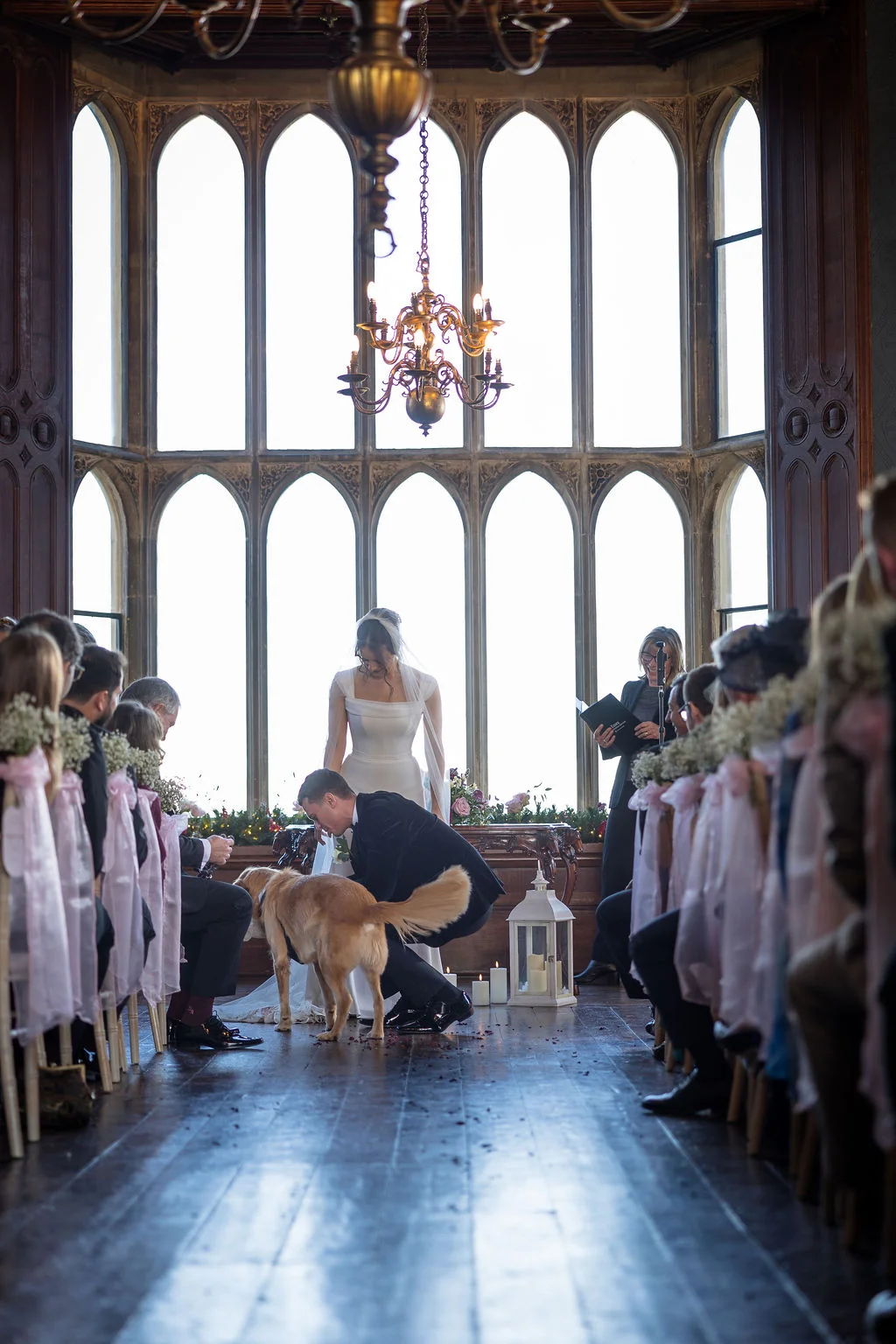A couple stands at the altar in a grand castle hall with tall arched windows, while their golden retriever dog joins them for the ceremony.