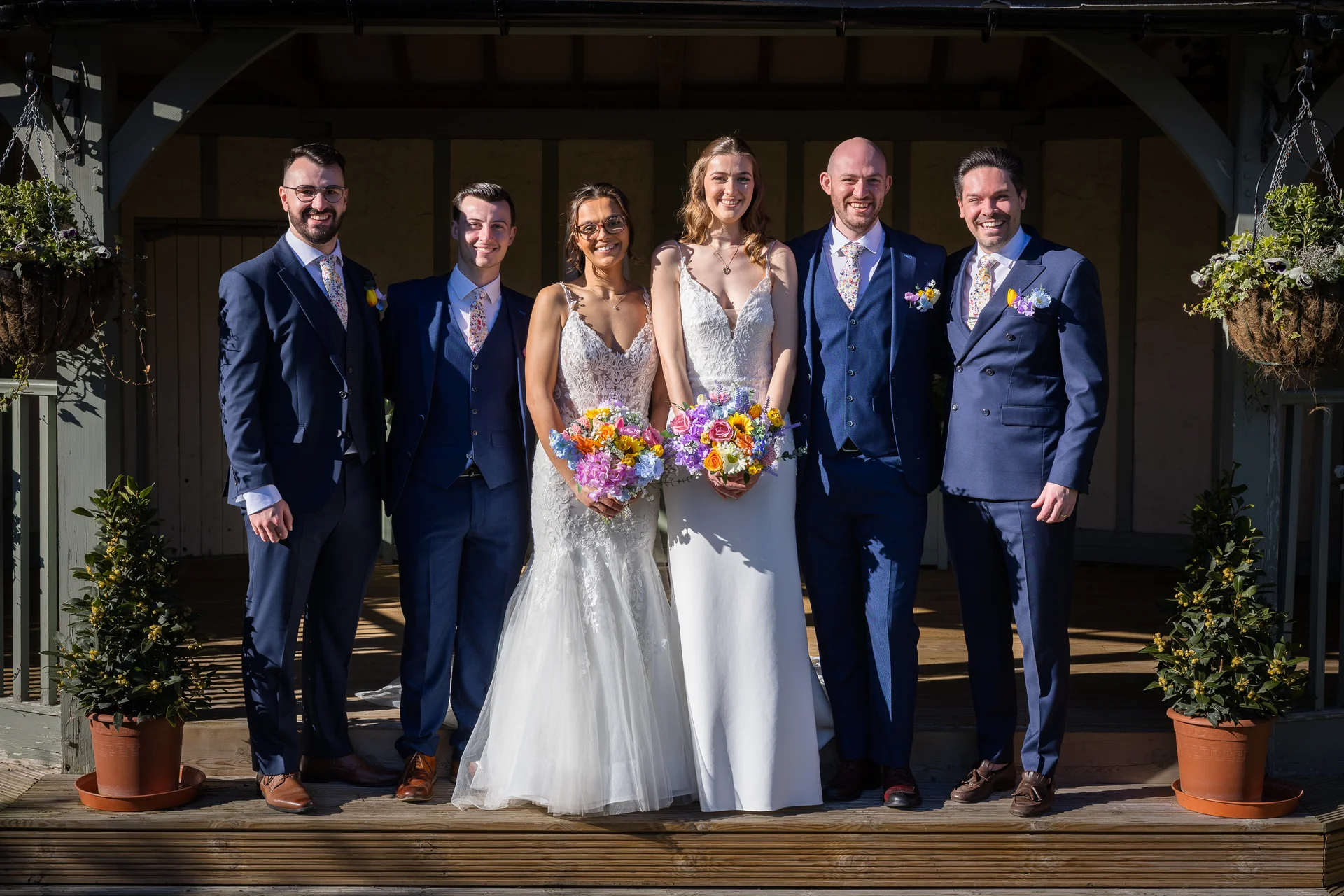 A smiling bridal party posing on a wooden deck in bright sunlight, featuring two brides in contrasting white gowns and four groomsmen in navy blue suits with colourful floral ties and vibrant bouquets.
