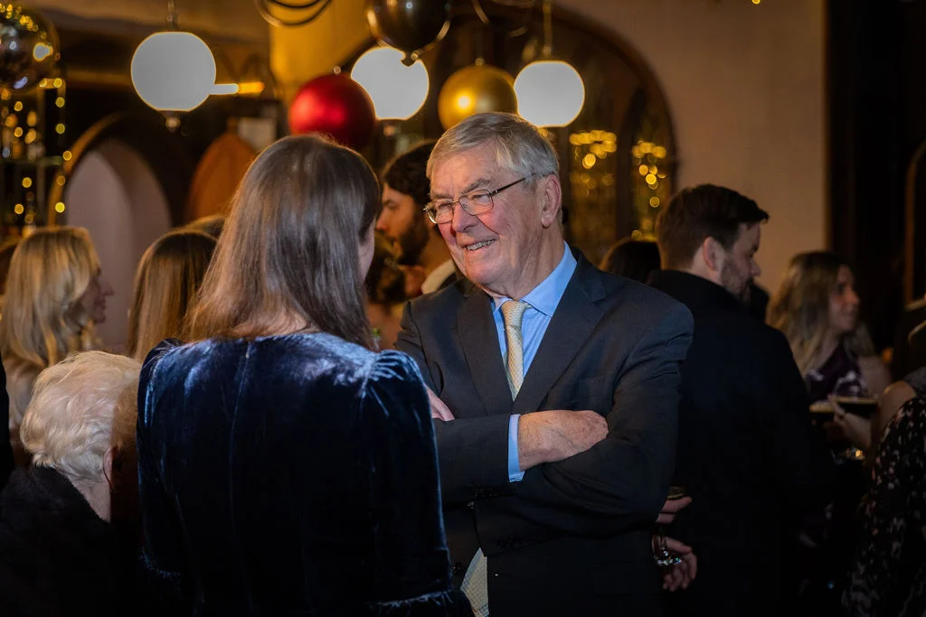 An elderly gentleman in a smart suit chats warmly with a guest during a cosy evening wedding reception illuminated by glowing orb lights.