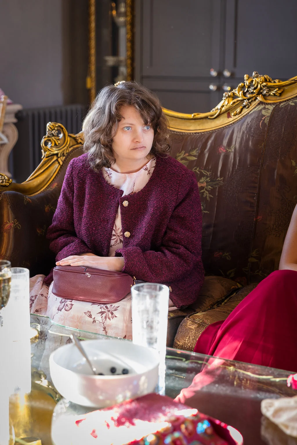 A wedding guest in a burgundy jacket and floral dress sits on an ornate gold-framed sofa during the reception.