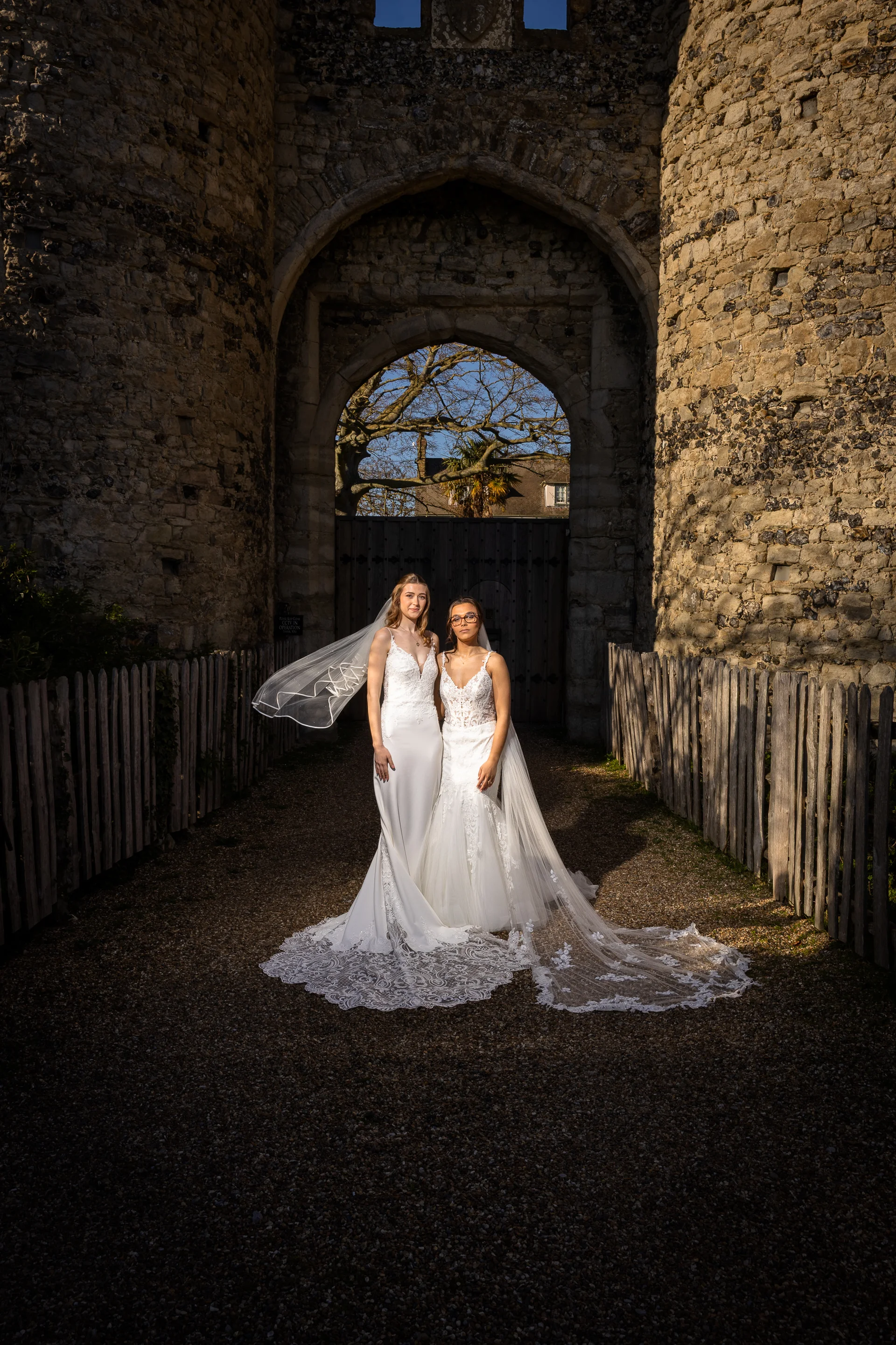 Two brides in elegant lace wedding gowns stand proudly framed beneath a majestic stone castle archway on a sunny day.