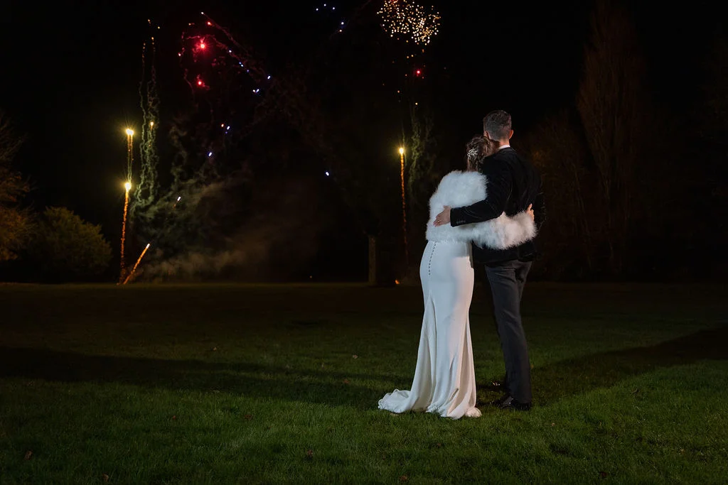 A newlywed couple embraces on a dark lawn at night, watching a vibrant fireworks display light up the sky.