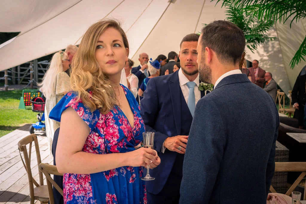 A wedding guest in a blue and pink floral dress holds a champagne flute while mingling under a stretch tent during the drinks reception.