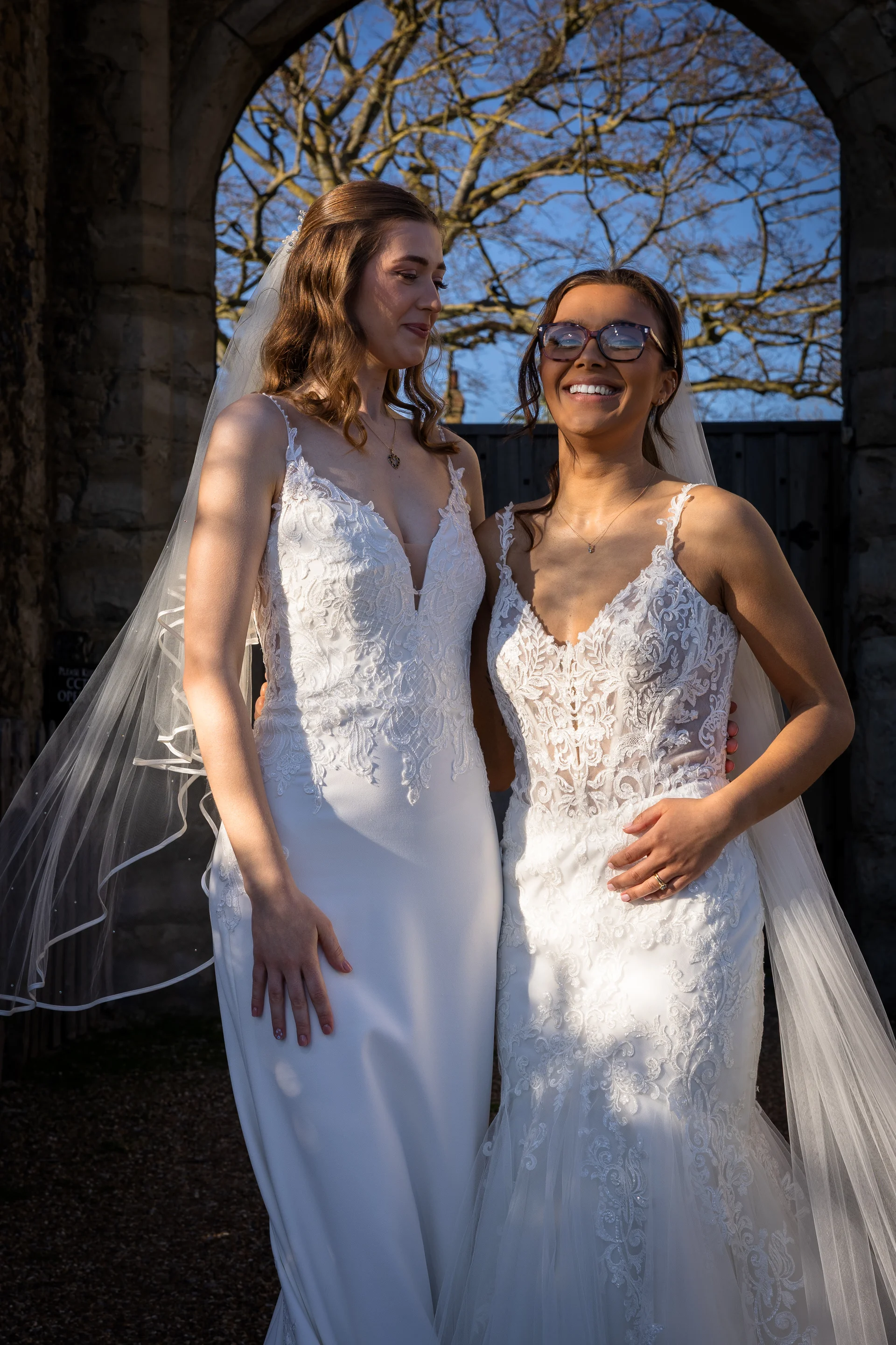 Two smiling brides pose together outdoors beneath a stone archway on a bright sunny day, wearing elegant lace wedding dresses and veils.