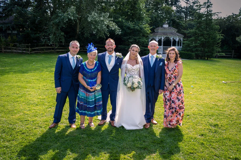 The bride and groom pose happily on a sunlit lawn with family members and groomsmen, with a white gazebo in the background.