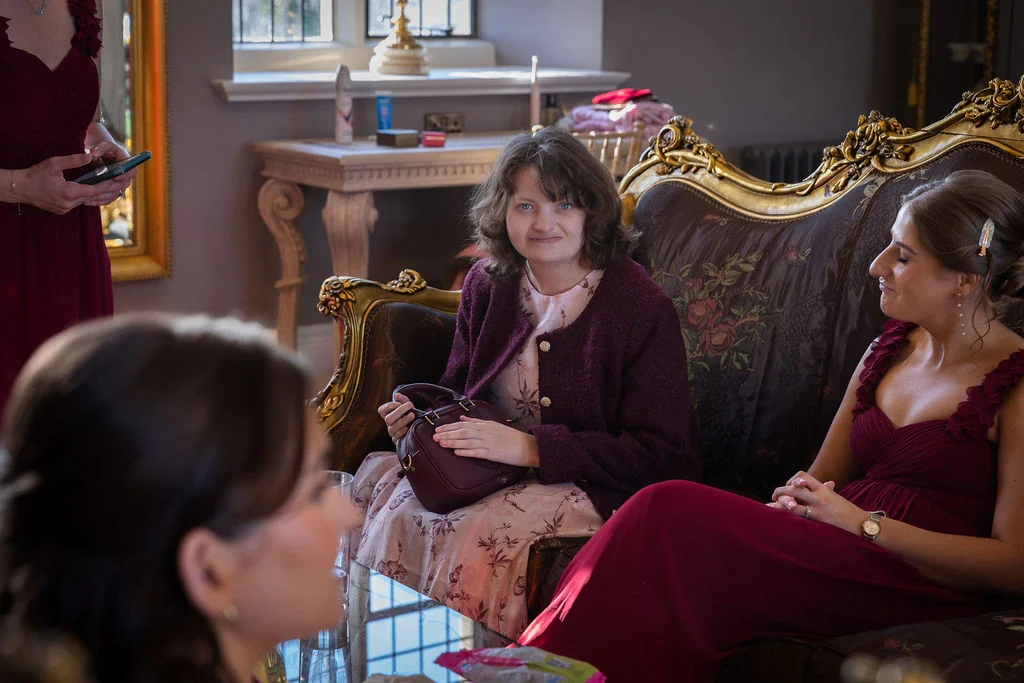 A candid indoor wedding shot capturing guests and bridesmaids relaxing and conversing on a vintage ornate sofa inside a stately home.