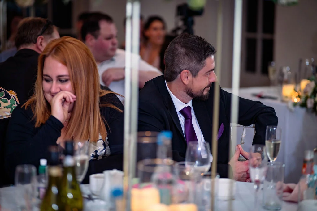 A guest in a dark suit and purple tie sits at a wedding breakfast table, smiling sideways while a woman with red hair sits next to him laughing.