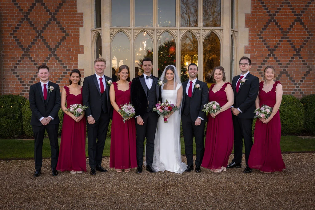 A bridal party dressed in black tuxedos with red ties and matching burgundy bridesmaid dresses poses in front of a red brick country house with large arched windows.