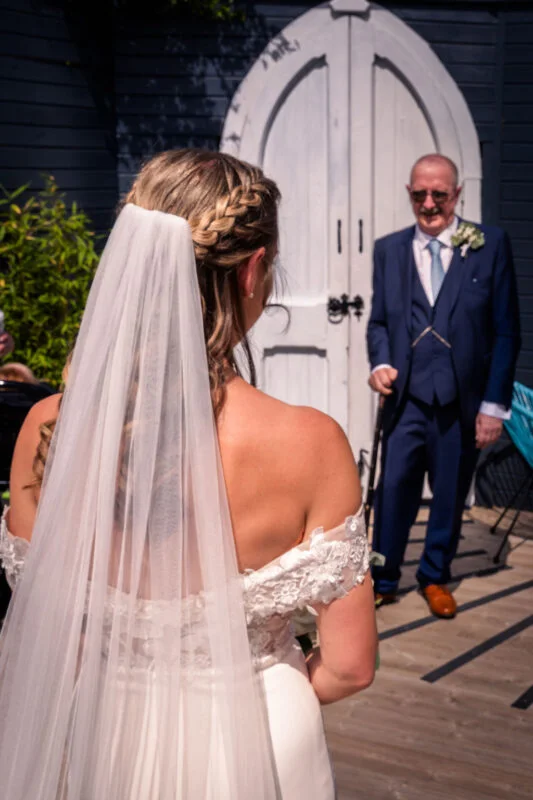 A bride in an off-the-shoulder gown and veil shares a touching first look moment with her smiling father in a navy suit outside white arched doors.