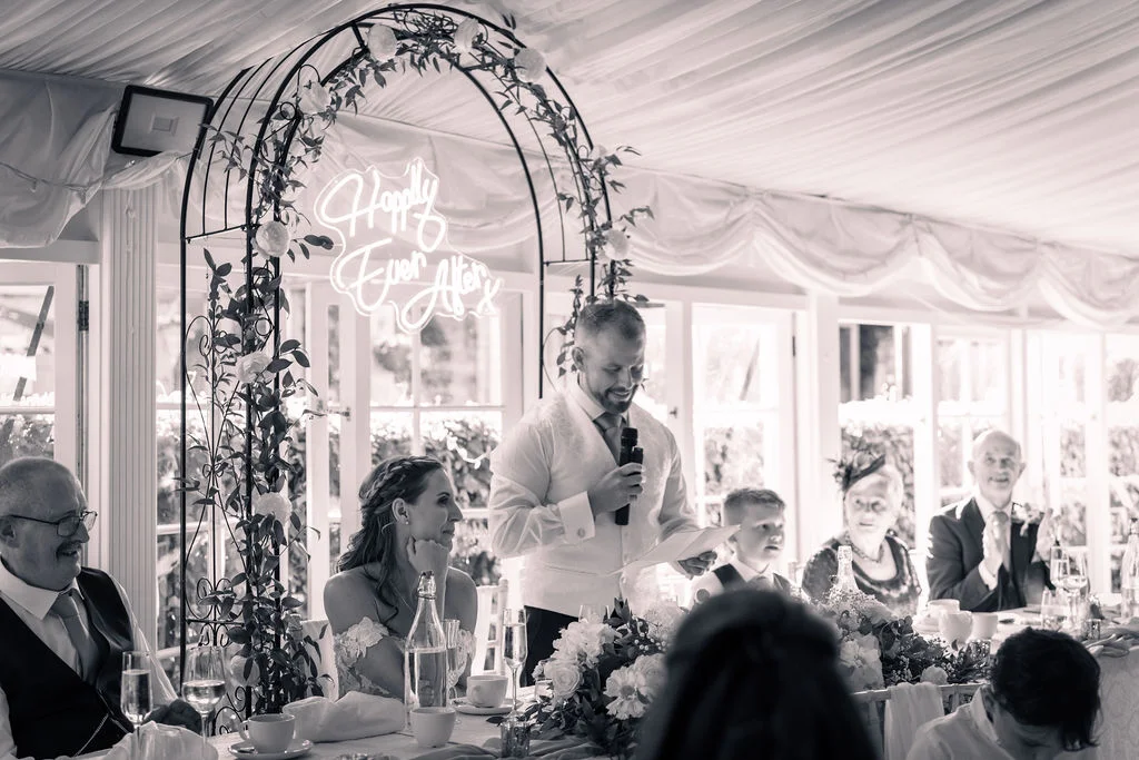 The groom delivers his speech at the top table under a floral arch featuring a neon sign inside a draped marquee.