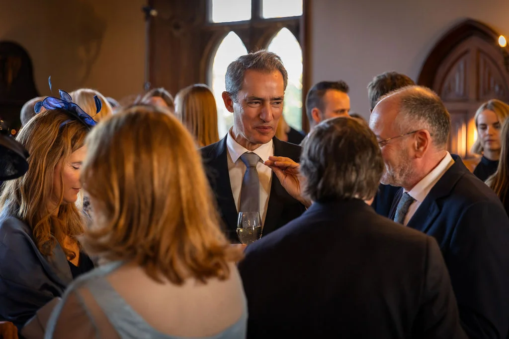Wedding guests mingle and converse during a drinks reception inside a historic venue with arched windows.