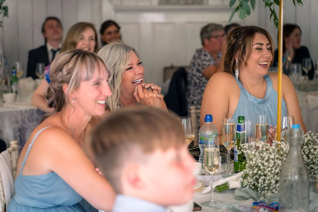 A group of smiling wedding guests laugh joyfully during the speeches at a reception table adorned with baby's breath floral arrangements.