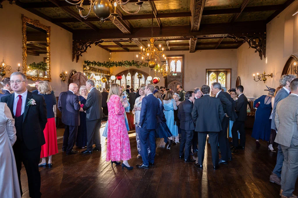 Wedding guests mingle during a festive drinks reception inside a grand, panelled country house reception room featuring a vaulted wooden ceiling and chandeliers.