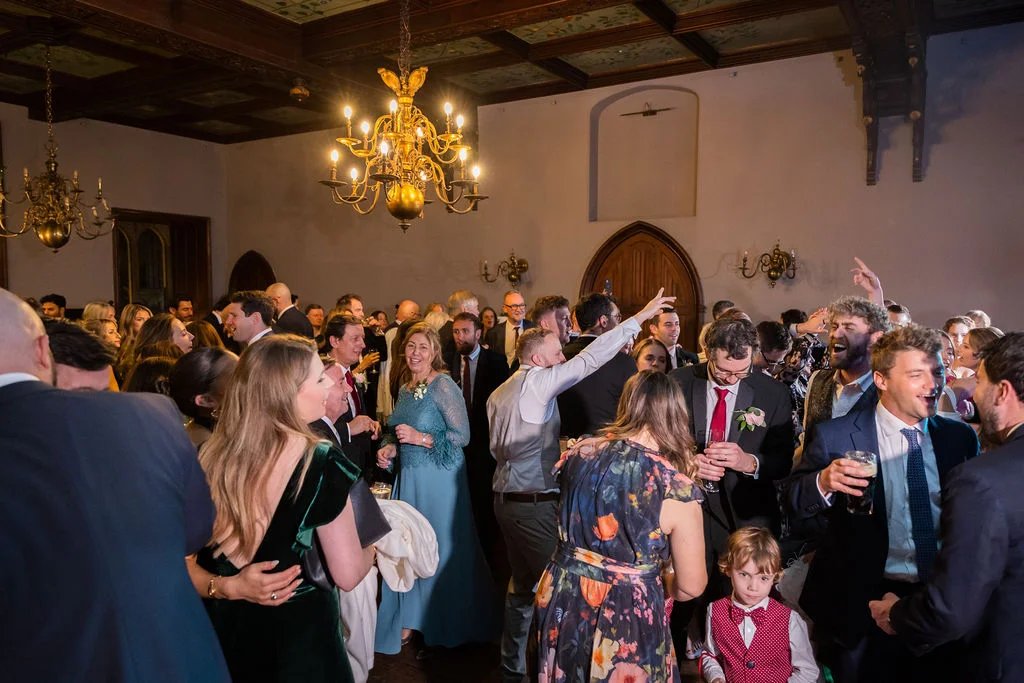 A lively wedding reception is underway inside a grand country house hall lit by ornate chandeliers, with guests socialising and celebrating together.