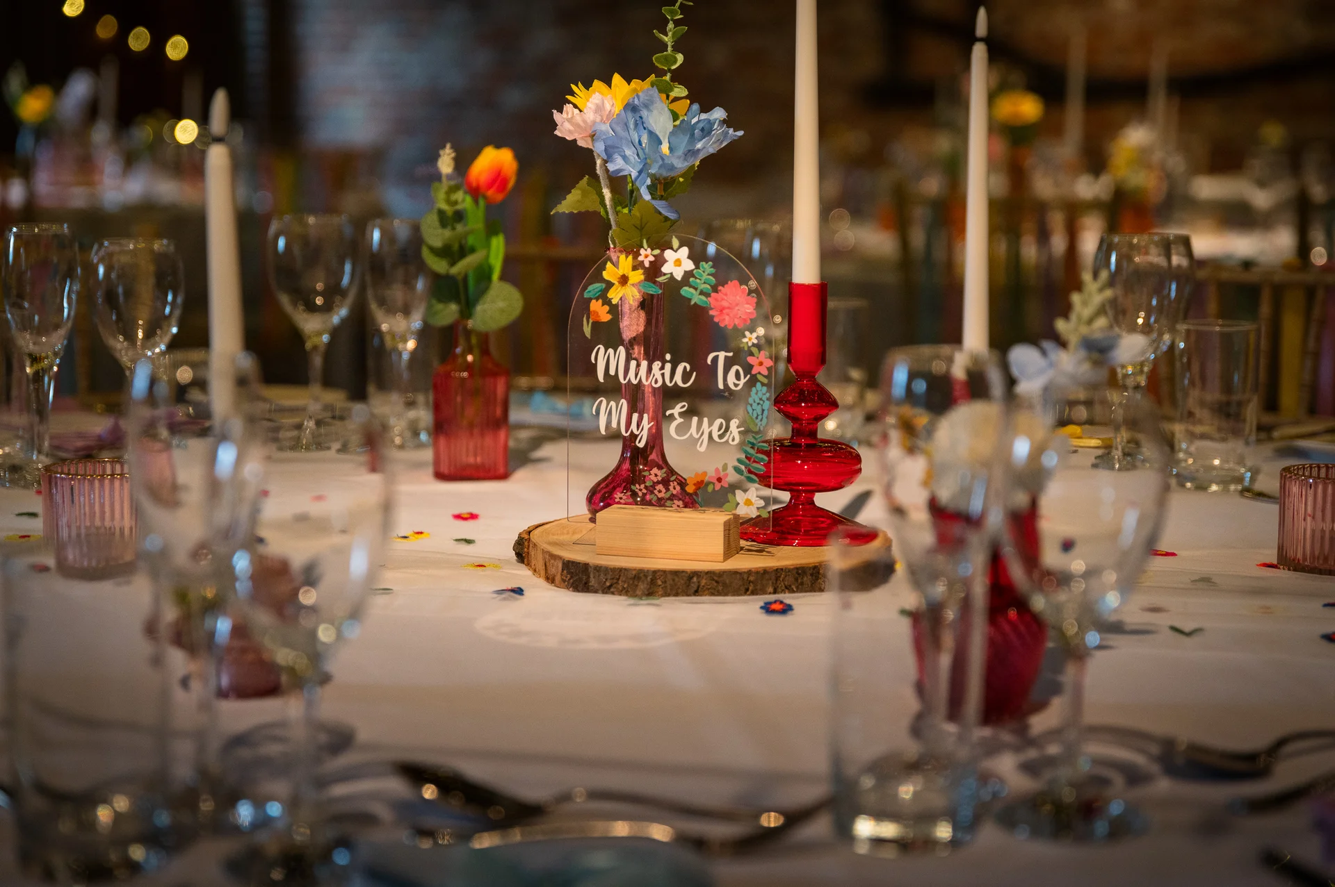 A vibrant wedding tablescape features colourful glass bud vases, whimsical wildflowers, a wooden centrepiece displaying an acrylic sign, and glowing taper candles.