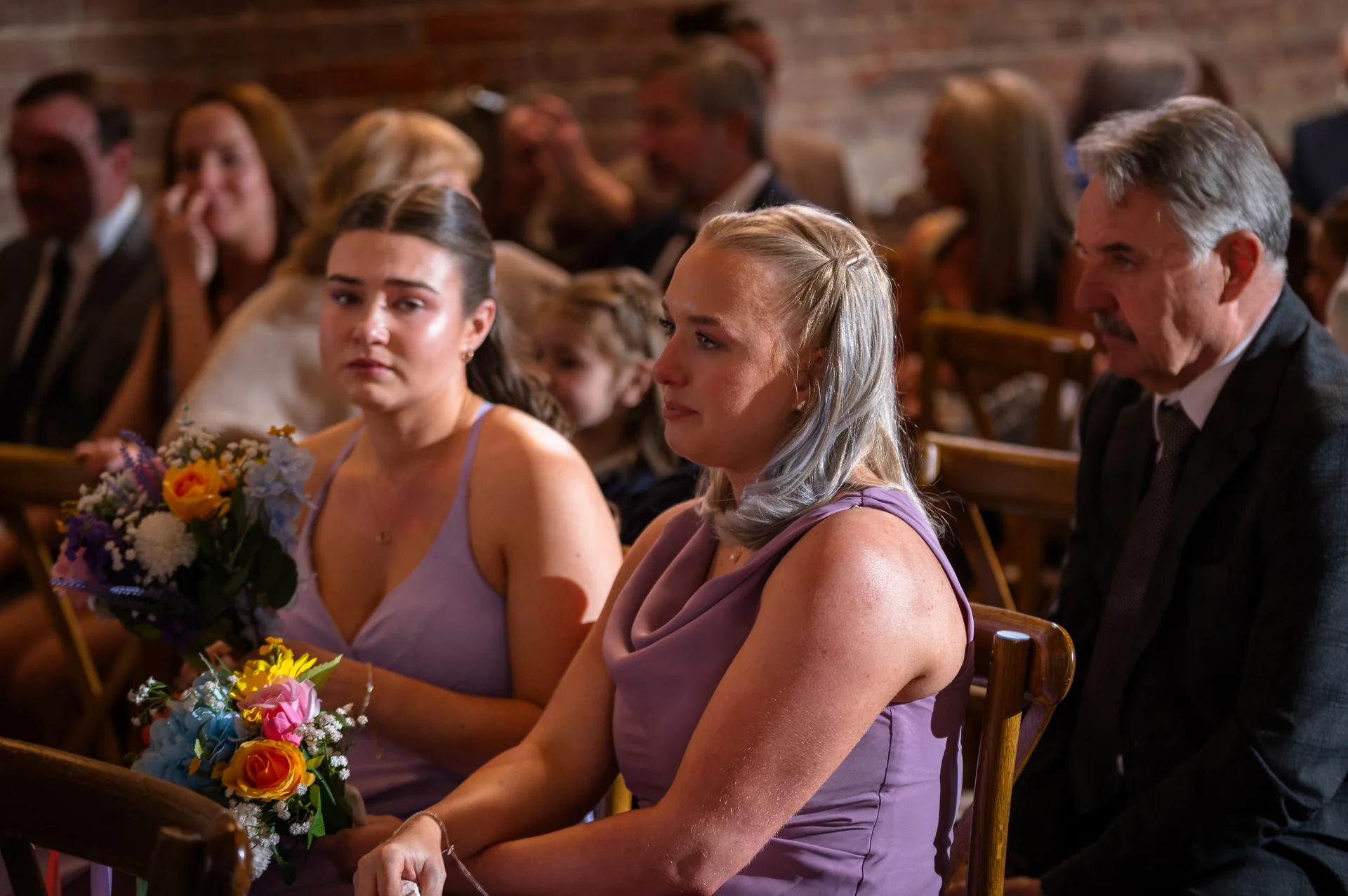 Emotional guests and bridesmaids holding colourful bouquets watch a wedding ceremony inside a venue with exposed brick walls.