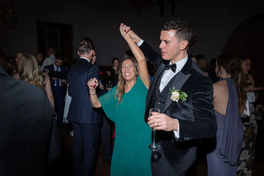A groom in a black velvet tuxedo dances joyfully on the dance floor with a guest wearing a green dress during the evening reception.