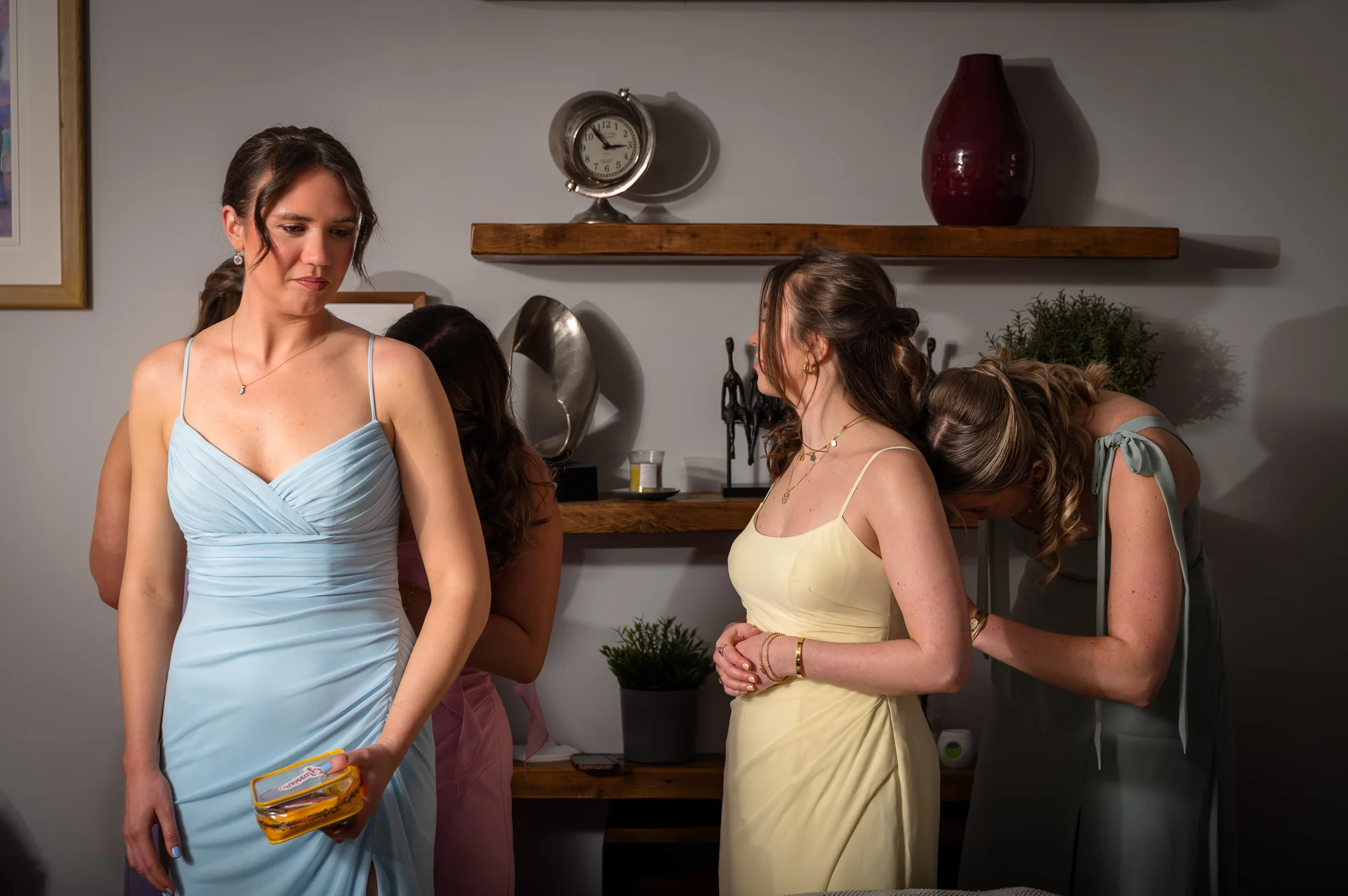 A group of bridesmaids wearing pastel dresses in various colours help each other with final preparations and styling indoors.