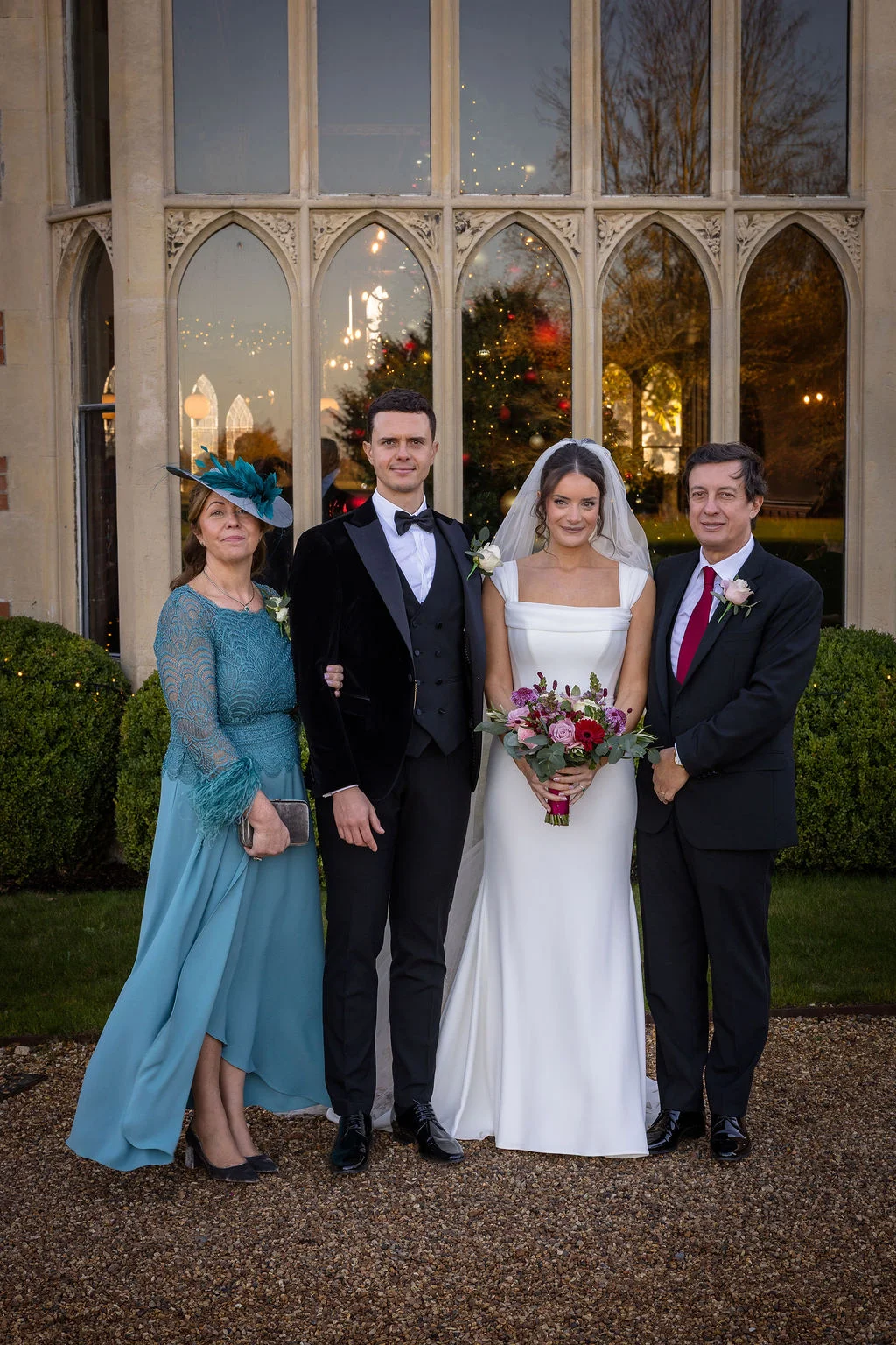 A newlywed couple poses outdoors with their parents in front of a grand stone manor house window with festive decorations visible inside.