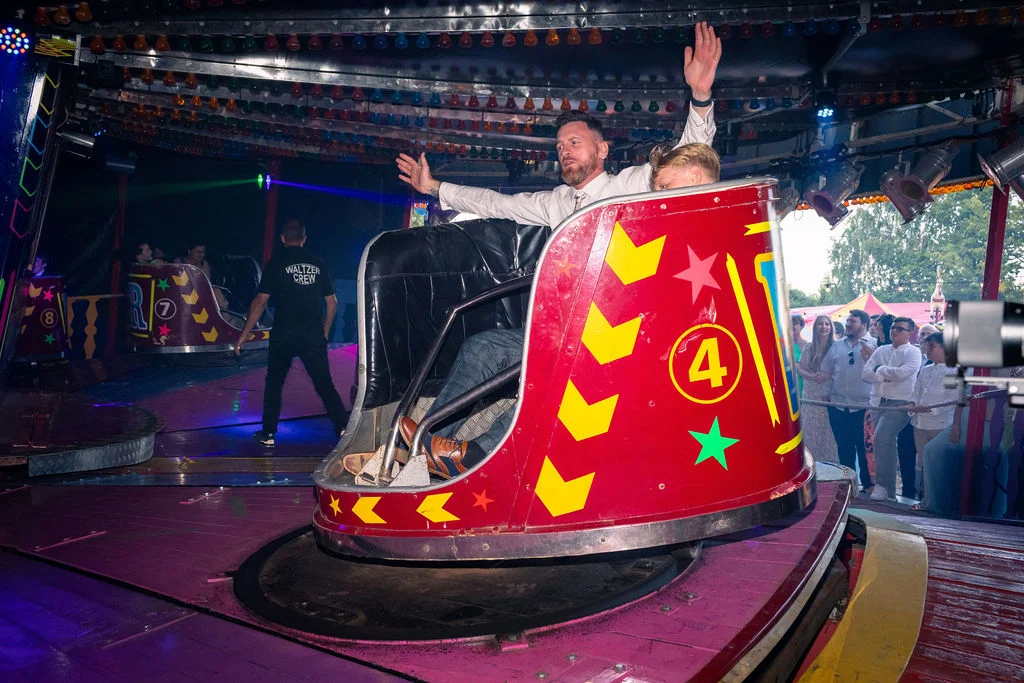 A groom and guest enjoy a ride on a bright red fairground waltz ride with colourful lights and a festival atmosphere during a wedding reception.