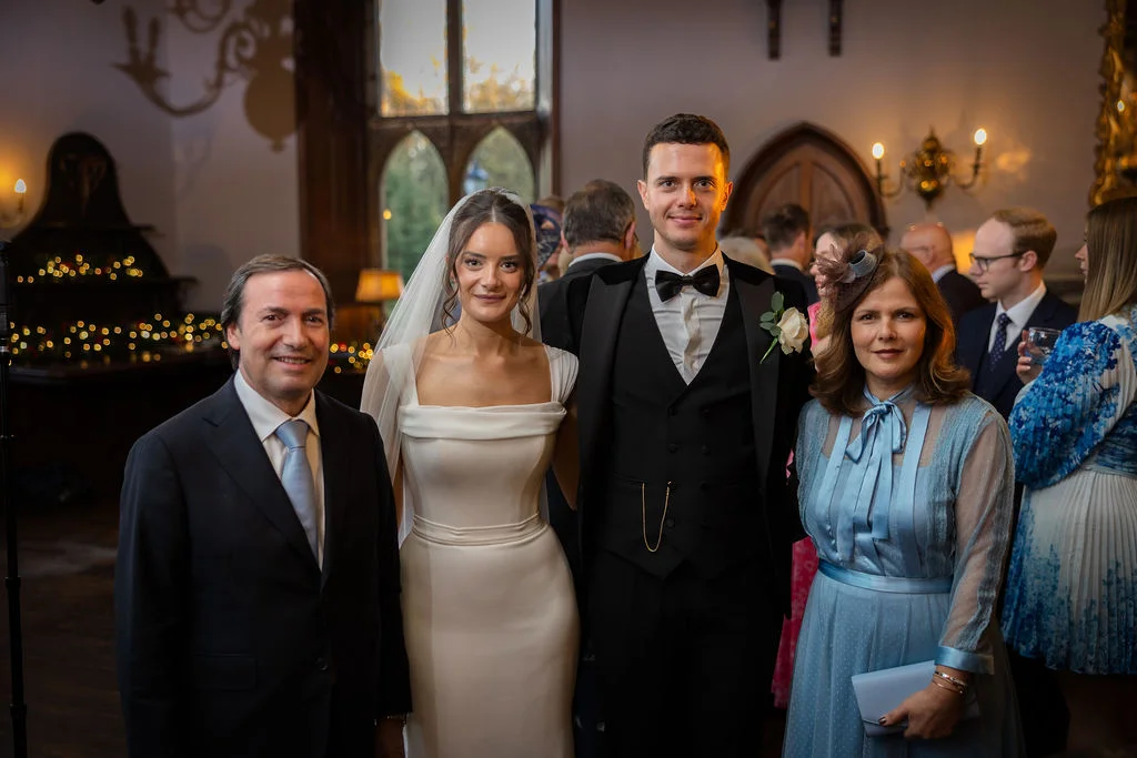 A newlywed bride in a sleek satin gown and veil stands alongside her groom in a black tuxedo, posing with family members inside an elegant historic venue during the wedding reception.