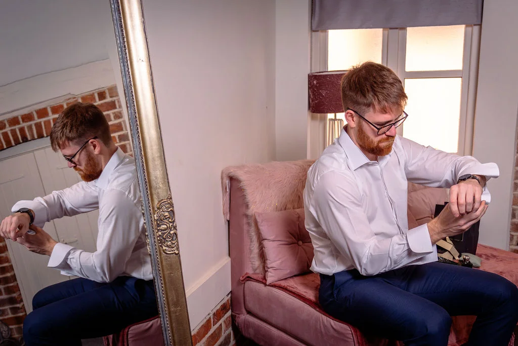 A groom sitting on a pink velvet chair adjusts his cufflinks while checking his watch, reflected in a large ornate mirror.