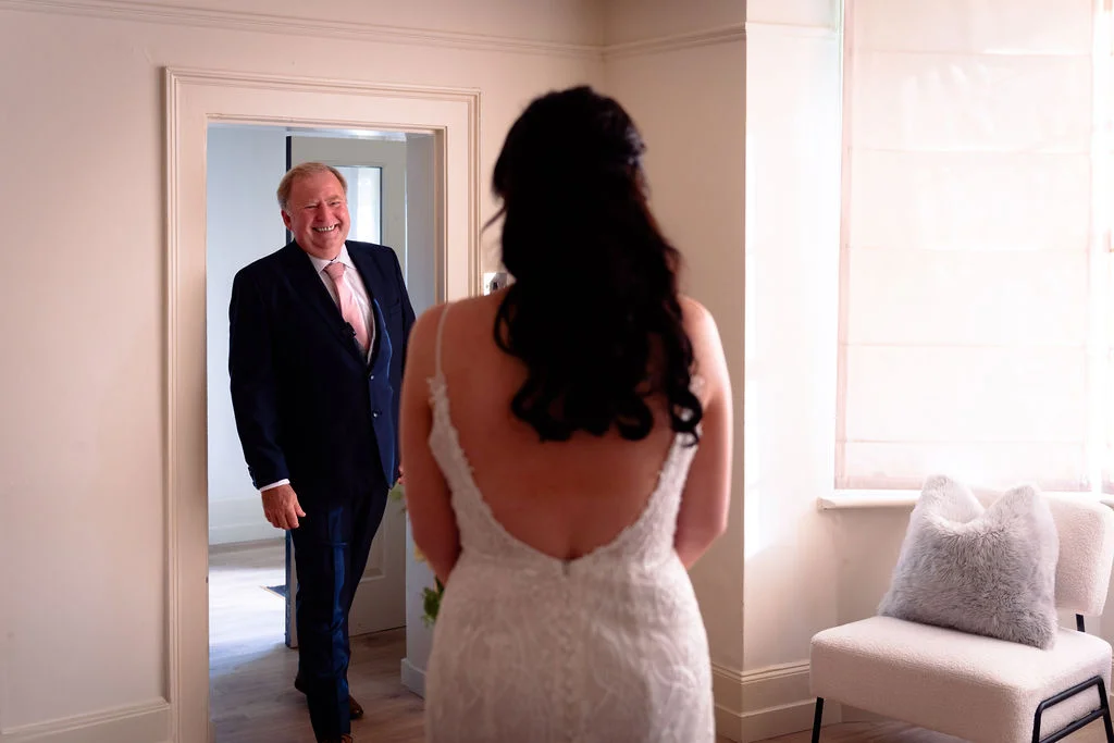 A smiling father of the bride reacts with pure joy during a first look moment, viewed from behind the bride in her wedding dress.