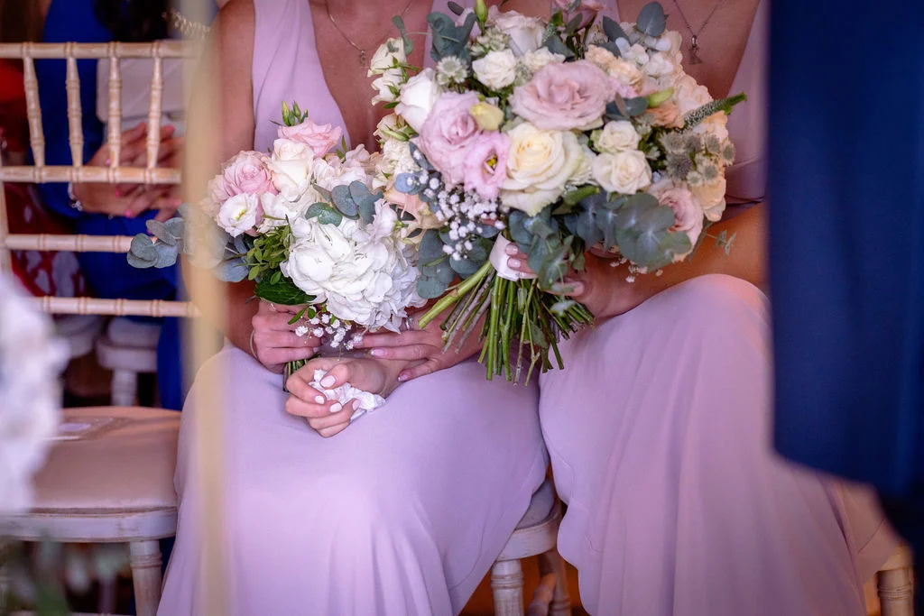 Two bridesmaids wearing matching pink chiffon dresses sit holding lush hand-tied bouquets featuring soft roses, hydrangeas, and eucalyptus foliage during the wedding ceremony.