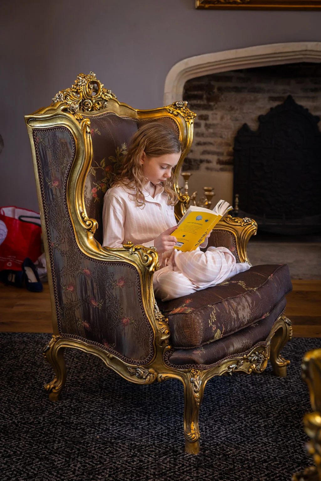 A young flower girl in matching pyjamas sits cross-legged reading a book in an ornate armchair indoors.