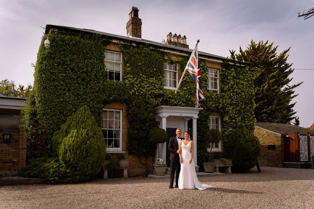 A bride and groom standing in front of a large house.
