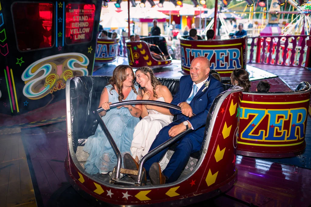 A bride, groom, and wedding guest laugh and smile while riding a waltzer fairground ride at a festival-themed wedding reception.
