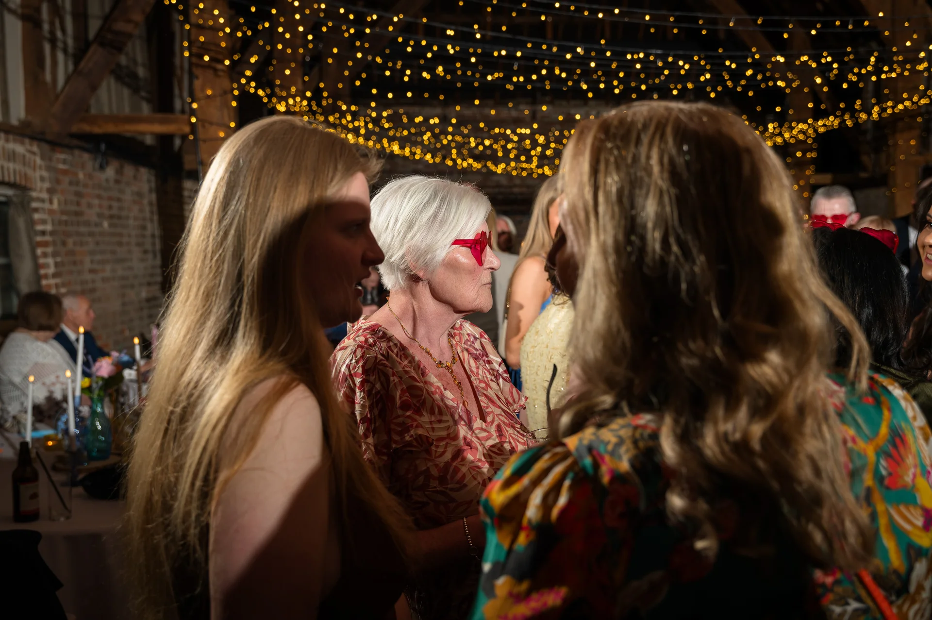 Wedding guests mingle and converse beneath a warm canopy of fairy lights inside a rustic barn reception venue.