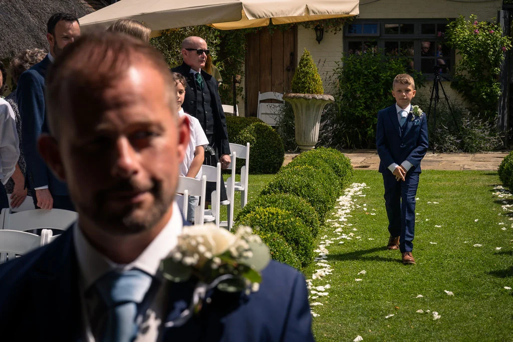 A young page boy in a smart navy suit walks down an outdoor grass aisle lined with petals, while a groom looks on from the foreground in soft focus.