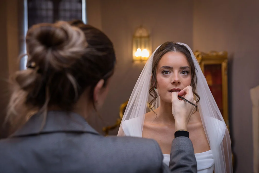 A bride wearing a veil and a satin gown has her makeup applied by a stylist during morning bridal preparations.