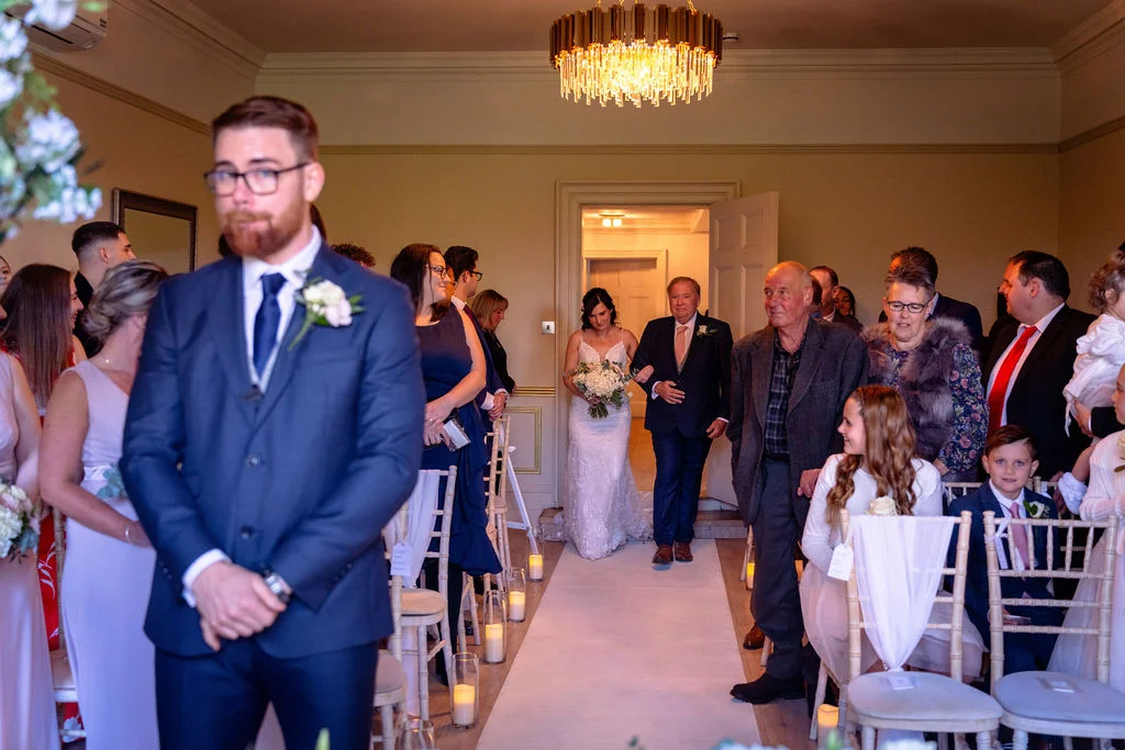 An emotional groom stands at the front of a light-filled indoor ceremony room while his bride walks down the aisle accompanied by her father.