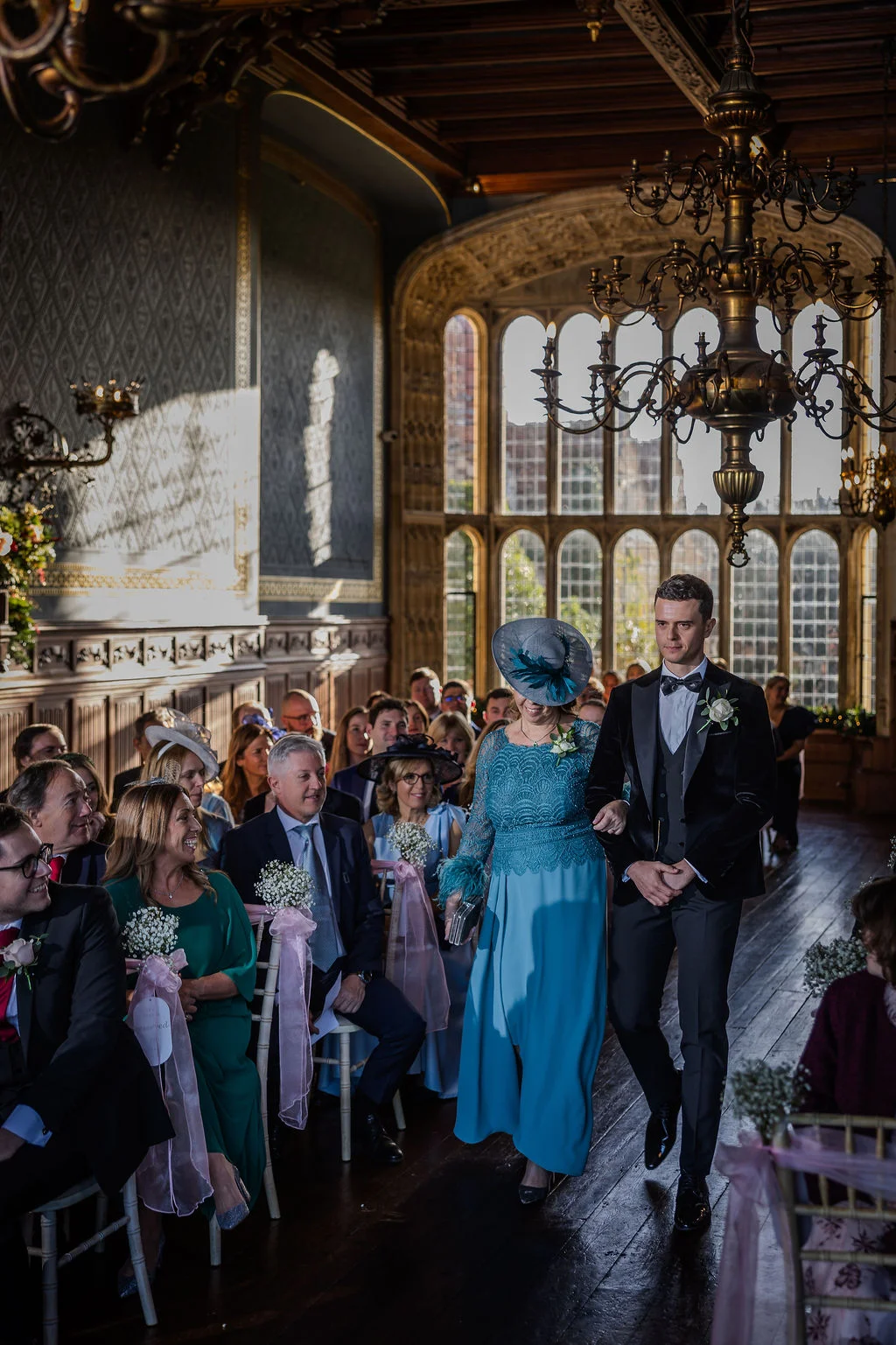 A groom in a black tuxedo and his mother walk down the aisle together during a grand country house wedding ceremony.