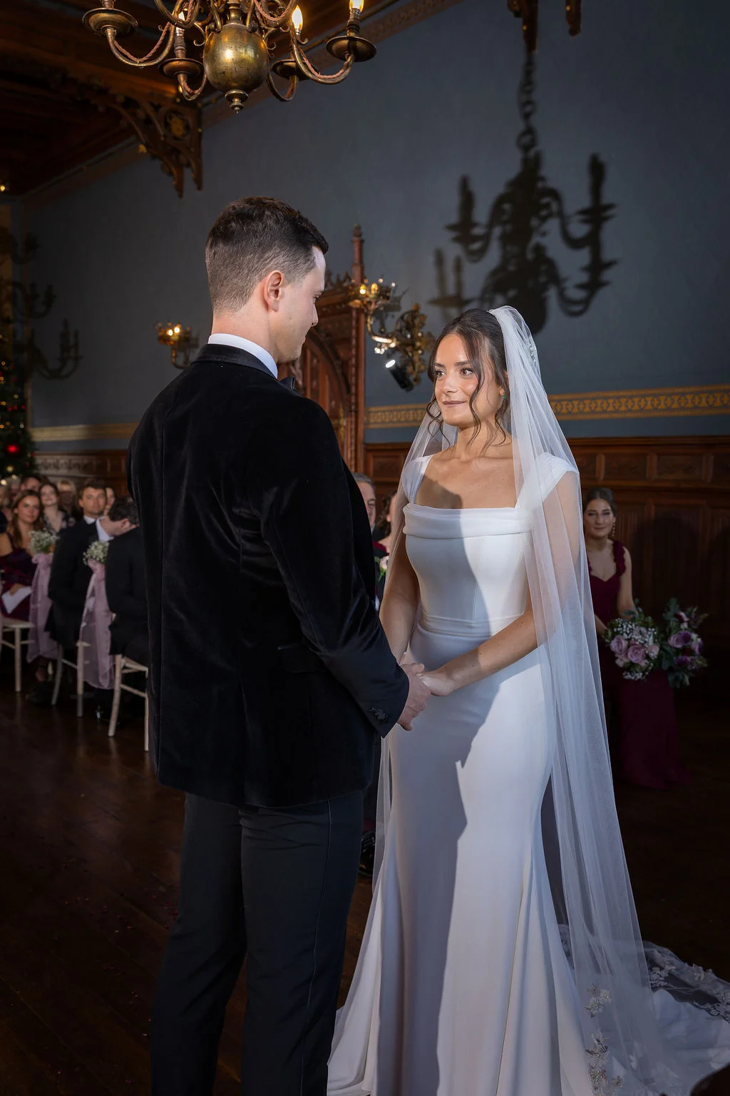 A groom in a black velvet tuxedo and a bride in a square-neck crepe gown with a long veil hold hands during their ceremony in a wood-panelled hall.