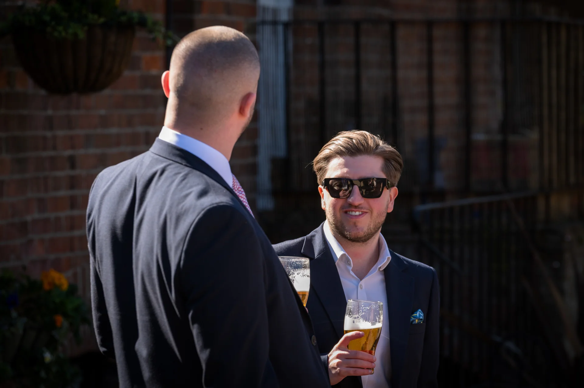 Two well-dressed guests in dark suits hold pints of beer and chat outdoors during a sunny wedding drinks reception.