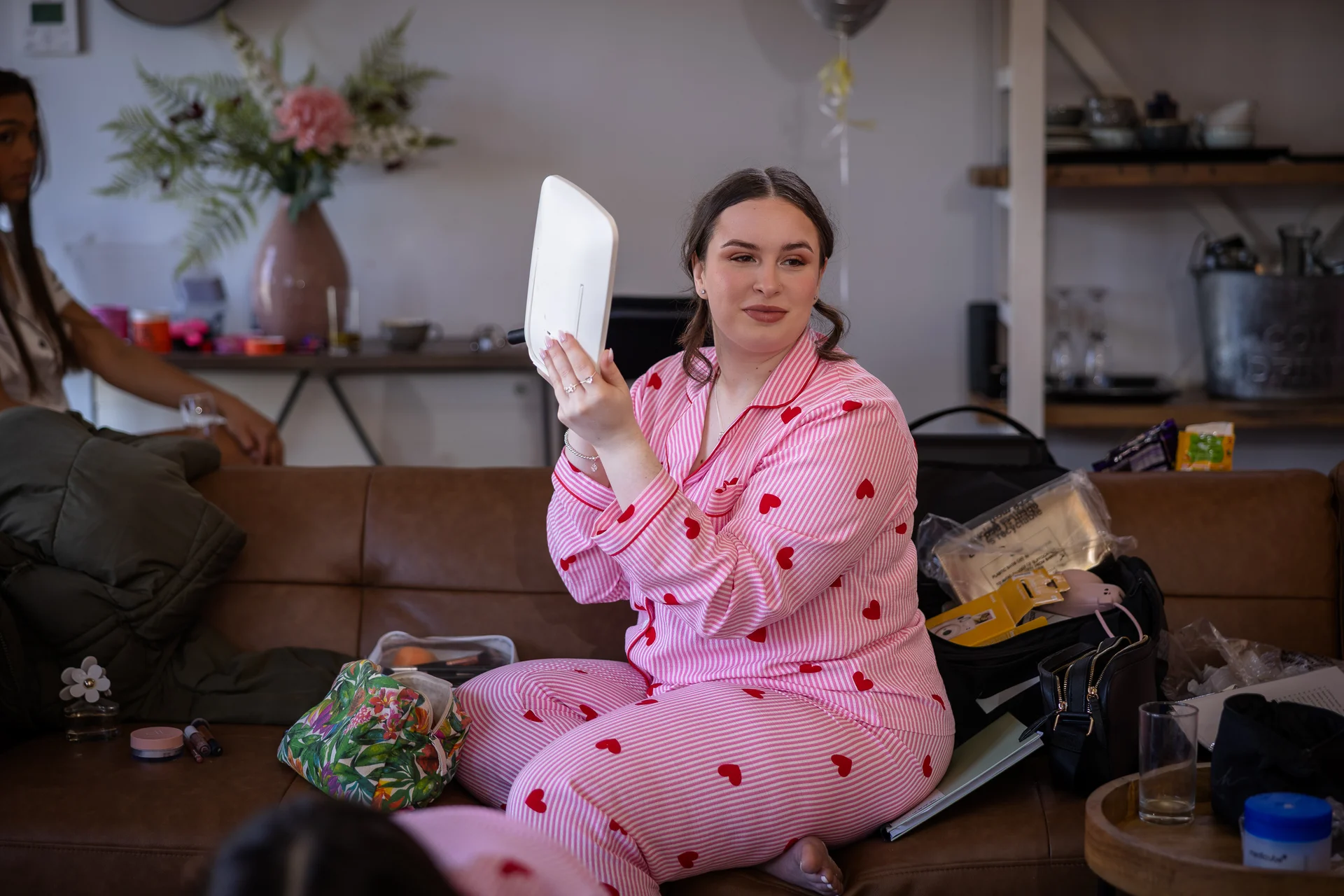A smiling bride wearing pink heart-printed pyjamas checks her makeup in a small handheld mirror while sitting on a leather sofa during morning bridal preparations.