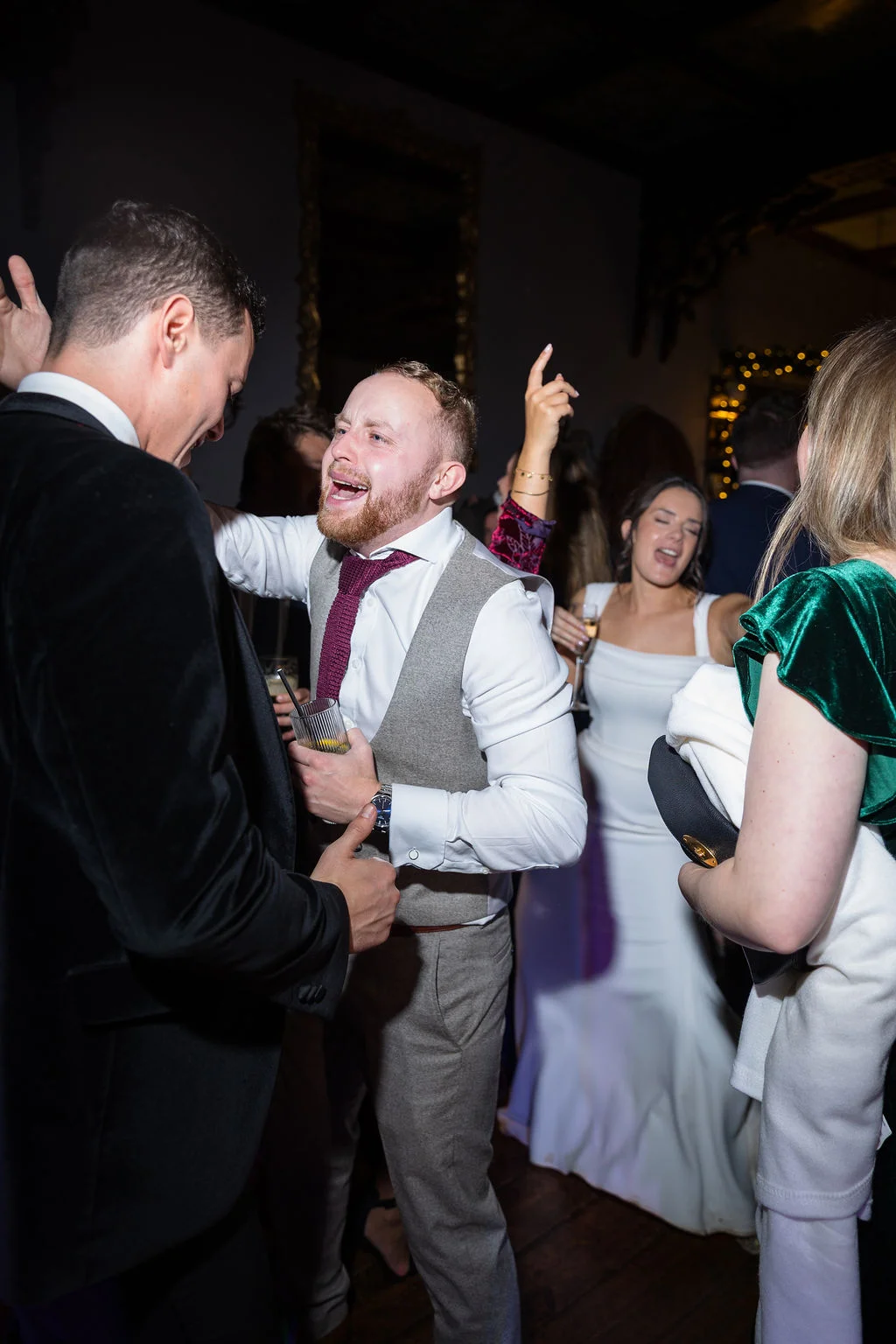 Wedding guests dancing and laughing together at an evening reception, with a groom in a grey waistcoat and patterned tie singing joyfully.