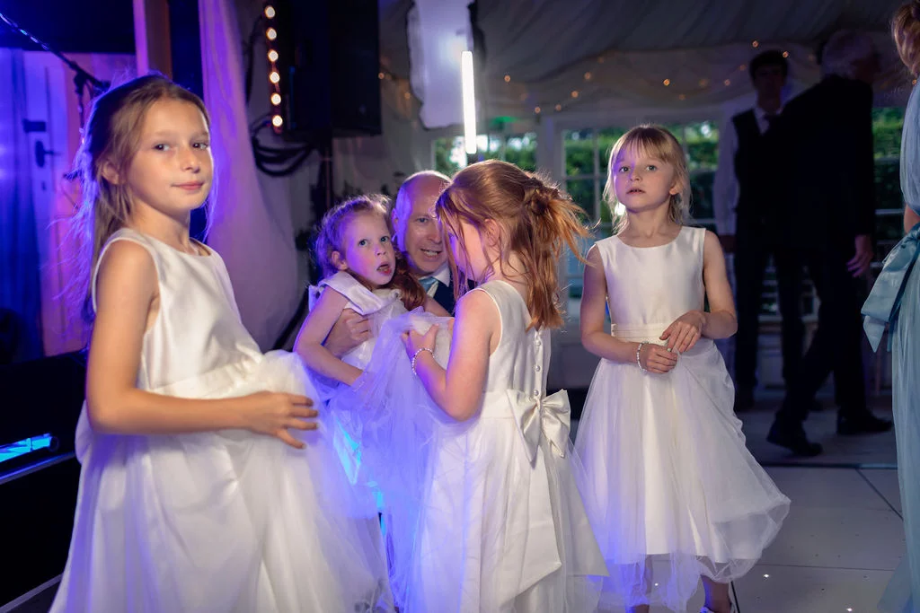 Several young flower girls in white dresses enjoy dancing on an illuminated dance floor during an evening wedding reception.