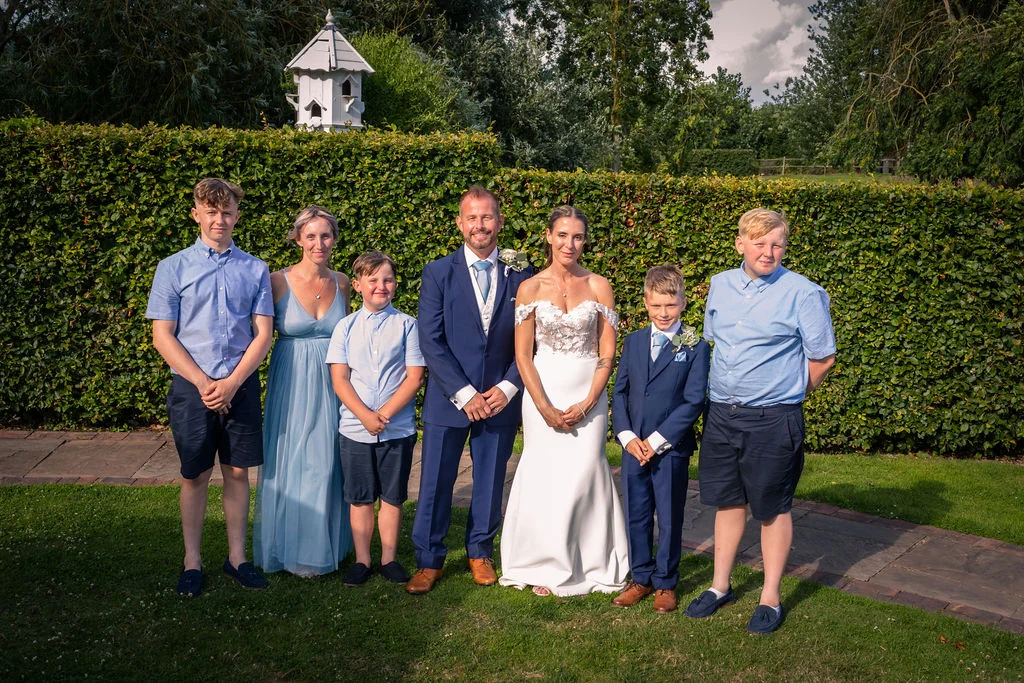 The newlywed couple stands proudly alongside their blended family in a manicured garden with a tall green hedge backdrop.