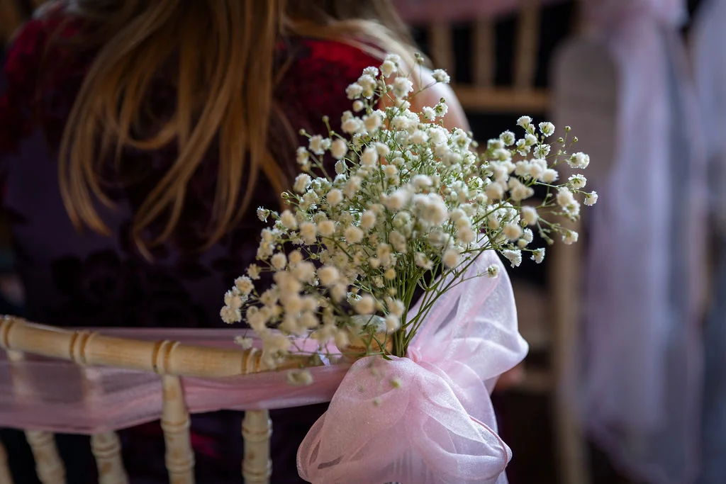 A close-up shot of a wooden wedding chair decorated with a pink organza sash and a delicate bunch of gypsophila flowers tied to the back.