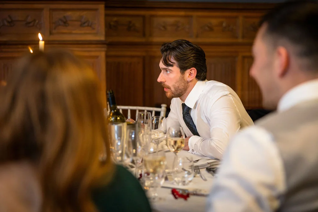 A wedding guest in a white shirt and dark tie listens intently during the speeches at a reception dinner in a wood-panelled room.