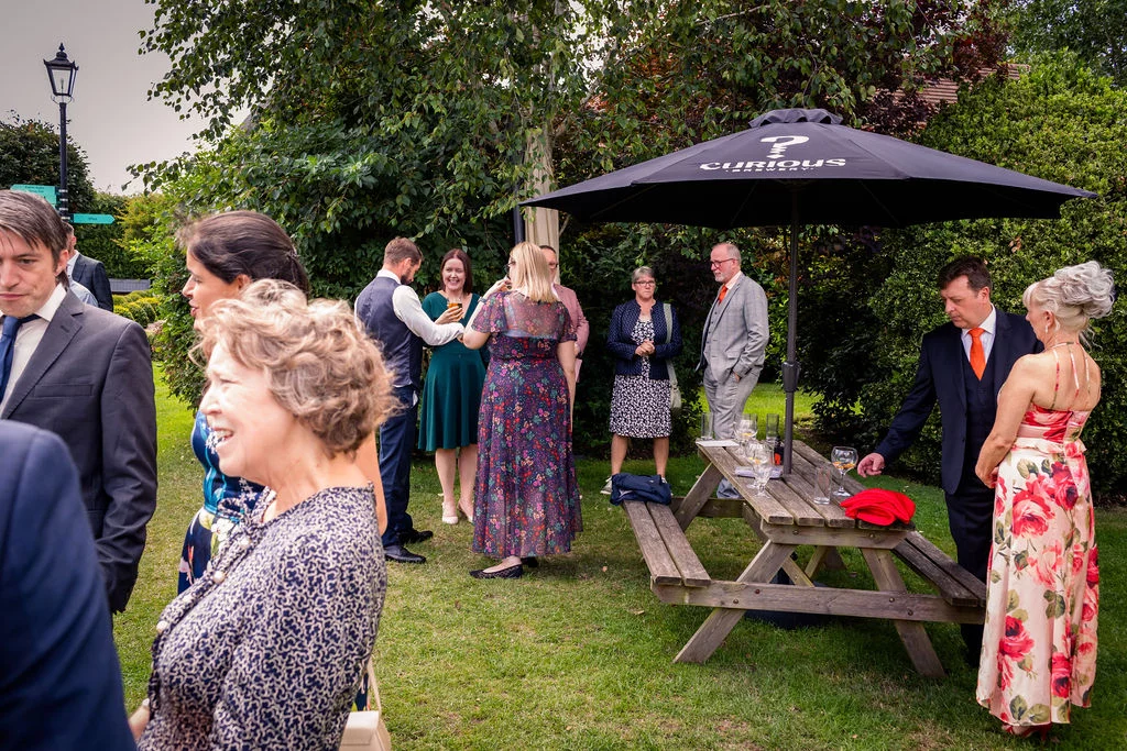 Wedding guests mingle and chat outdoors on a grassy pub garden during the drinks reception under an overcast sky.
