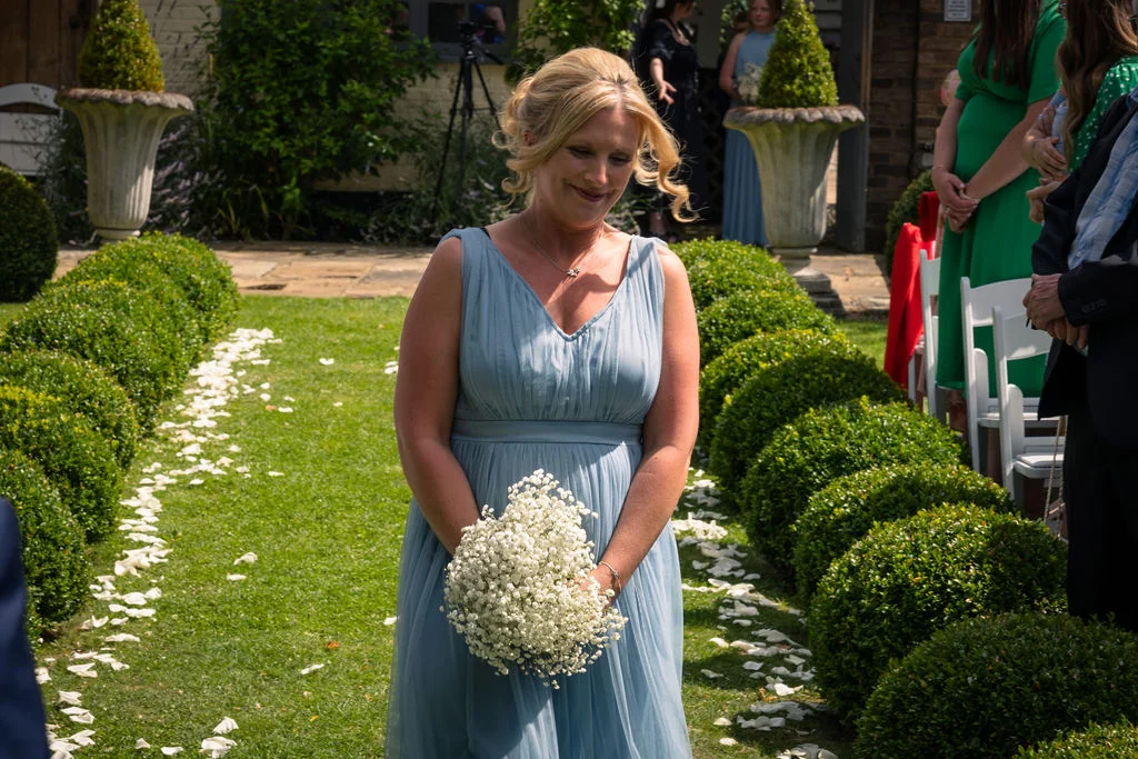 A bridesmaid in a dusty blue chiffon dress walks down a garden aisle lined with white petals and neat topiary bushes, carrying a bouquet of gypsophila.