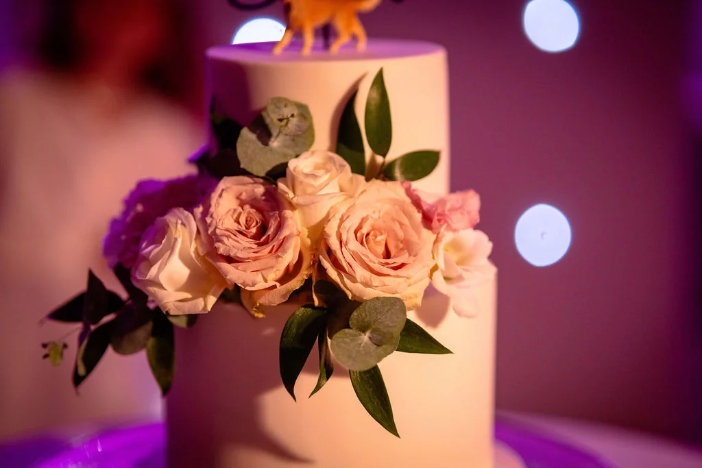 A close-up of a two-tier white fondant wedding cake decorated with fresh blush roses and eucalyptus leaves against a softly lit background.