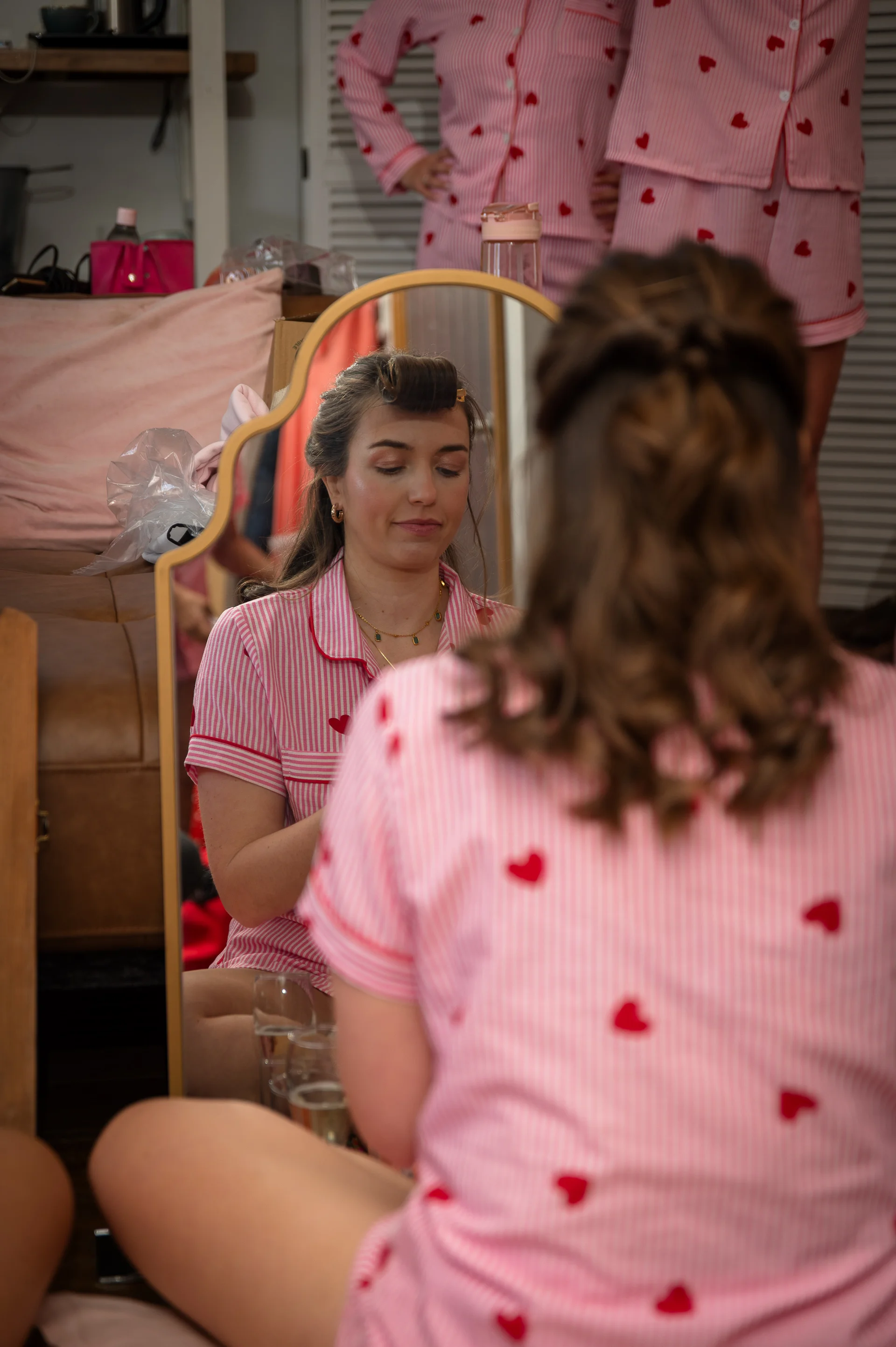A bride sits in front of a mirror during bridal prep, wearing matching pink and white heart-patterned pyjamas while styling her hair.