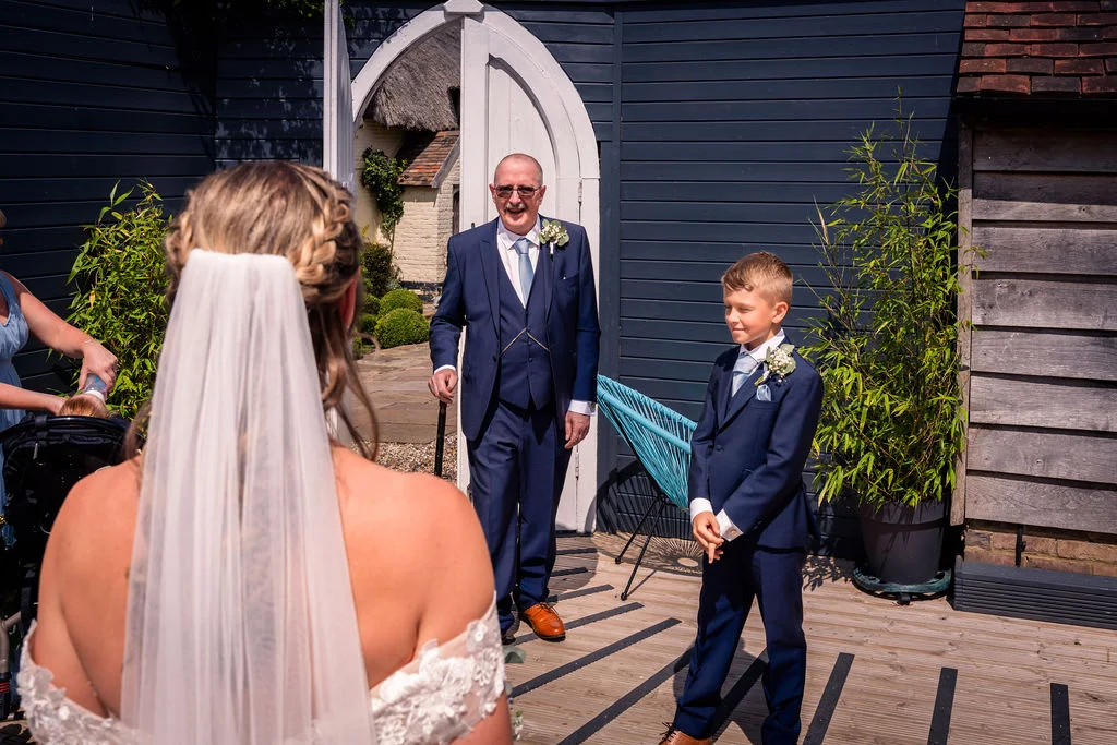A bride in a veil seen from behind looks towards her father and a young page boy standing outside in front of dark wooden cladding on a sunny day.